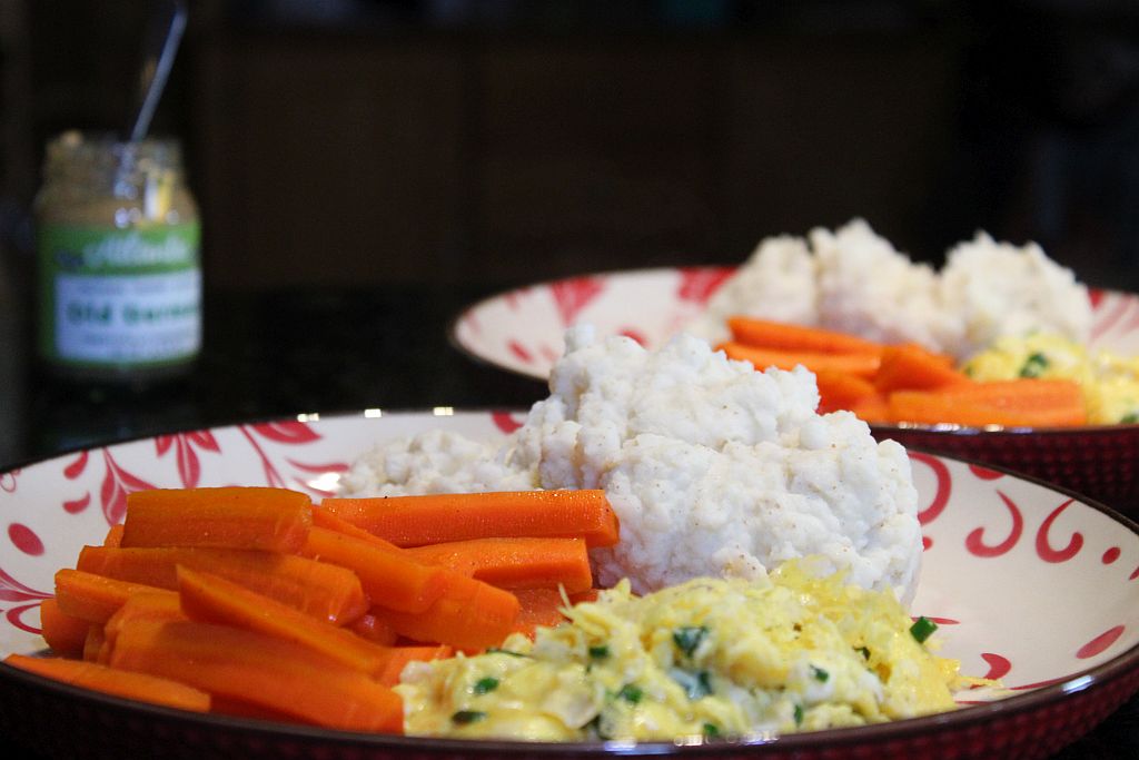 Two plates with mashed potatoes, carrots and scrambled eggs, a jar of mustard in the back